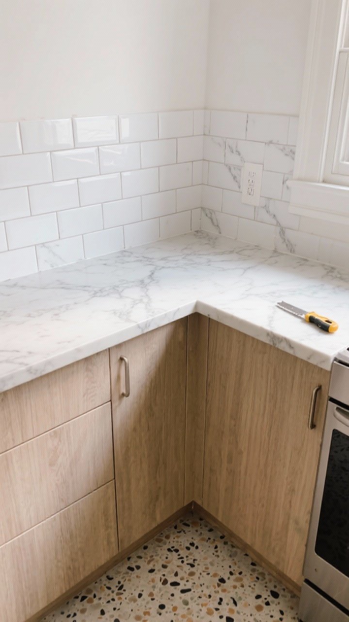 A bright, small rental kitchen corner with peel-and-stick upgrades: closeup detail shot of a classic white subway tile peel-and-stick backsplash meeting a marble-look vinyl countertop cover with subtle gray veining; matte wood-look peel-and-stick on flat cabinet fronts, and terrazzo-pattern peel-and-stick floor tiles underfoot. Warm natural daylight, clean trimmed edges, smooth surface finish, utility knife and smoothing tool resting to the side. Photorealistic, straight-on angle emphasizing textures and realism.