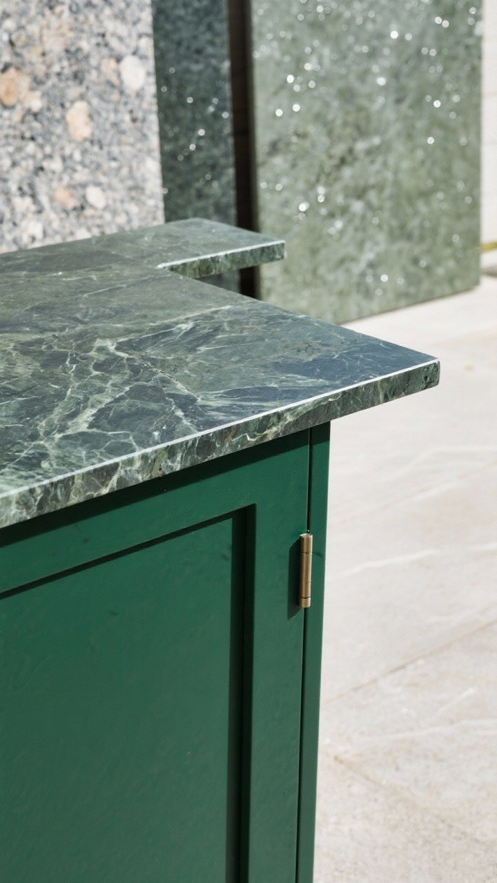 Closeup, raking-angle detail of outdoor countertop edges and finishes: a deep green base cabinet topped with velvety soapstone showing subtle veining, an eased edge profile, a small cantilevered bar ledge extension; adjacent sample boards of honed granite and sintered stone in green-gray tones; droplets of water beading to imply durability; neutral, even daylight to reveal surface texture without glare.